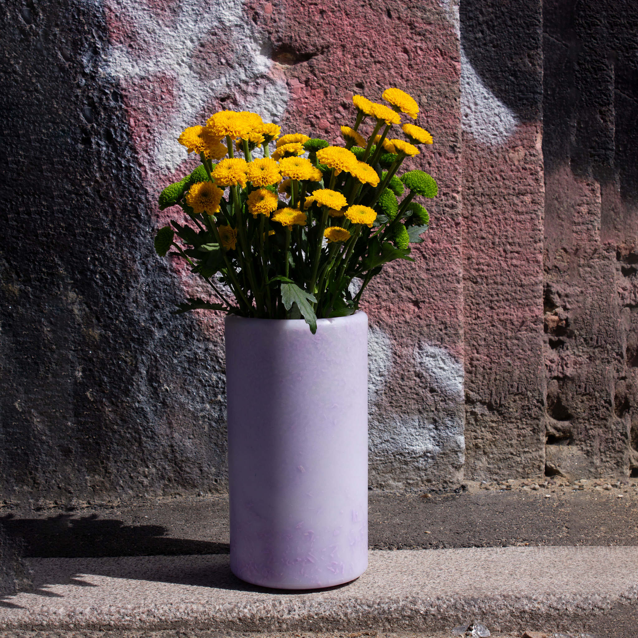Baby Donna Vase in purple by SMALLrevolution, made from recycled plastic waste and styled with yellow flowers against a textured graffiti wall. The image showcases Danish circular design that transforms discarded plastic into functional, long-lasting decor.