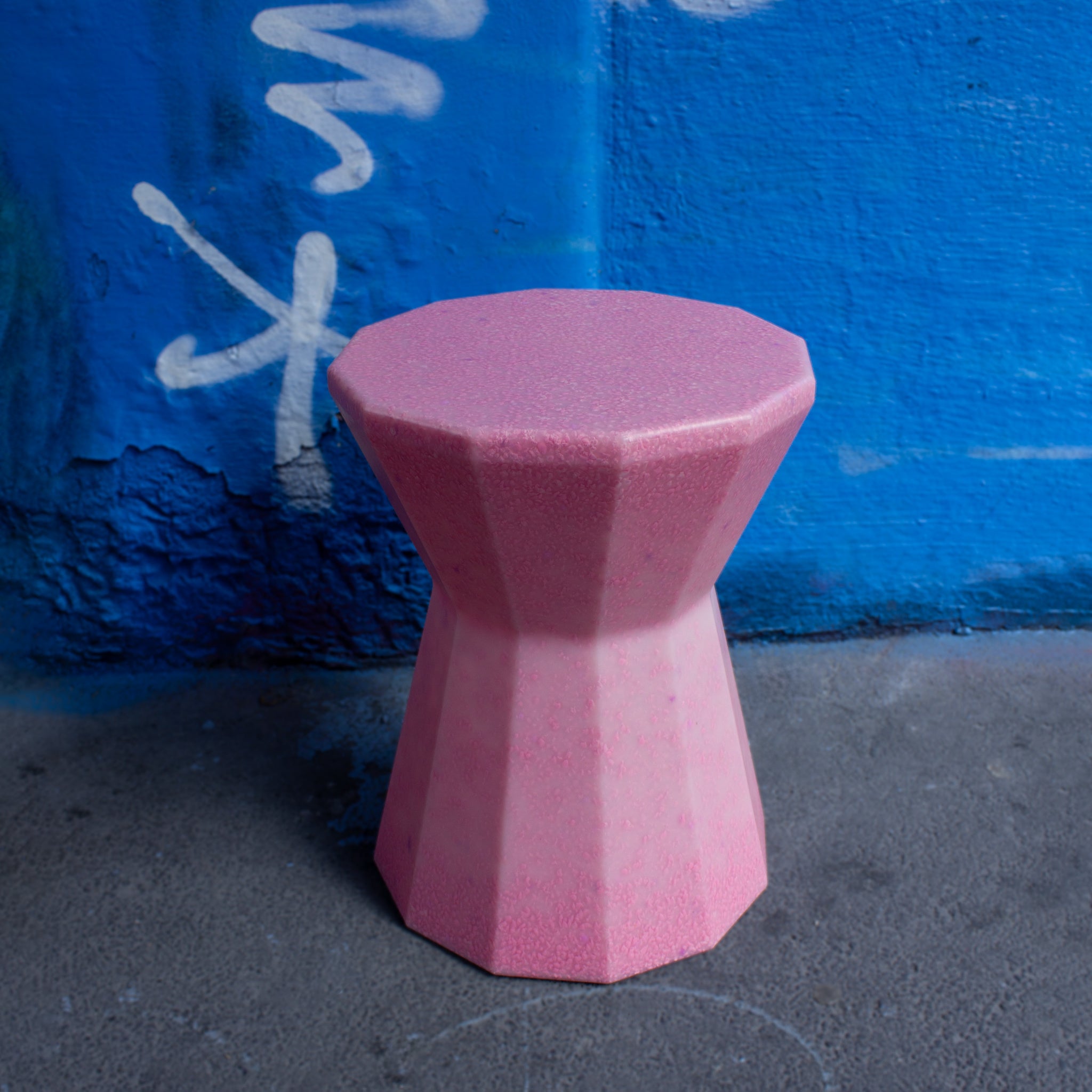 The Mabel Stool in pink by SMALLrevolution, made from 100% recycled plastic waste, photographed against a blue graffiti wall. Designed for both indoor and outdoor use, the stool showcases how waste materials can be transformed into durable, colorful furniture.