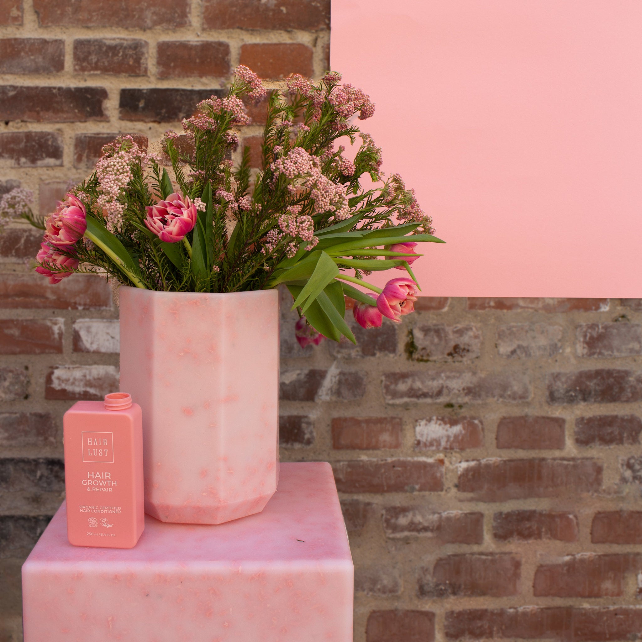 Pink Wangari Vase by SMALLrevolution made from recycled plastic waste, styled with pink flowers and Hairlust packaging against a brick wall. The image highlights Danish circular design that turns plastic waste into durable, decorative furniture and home objects.
